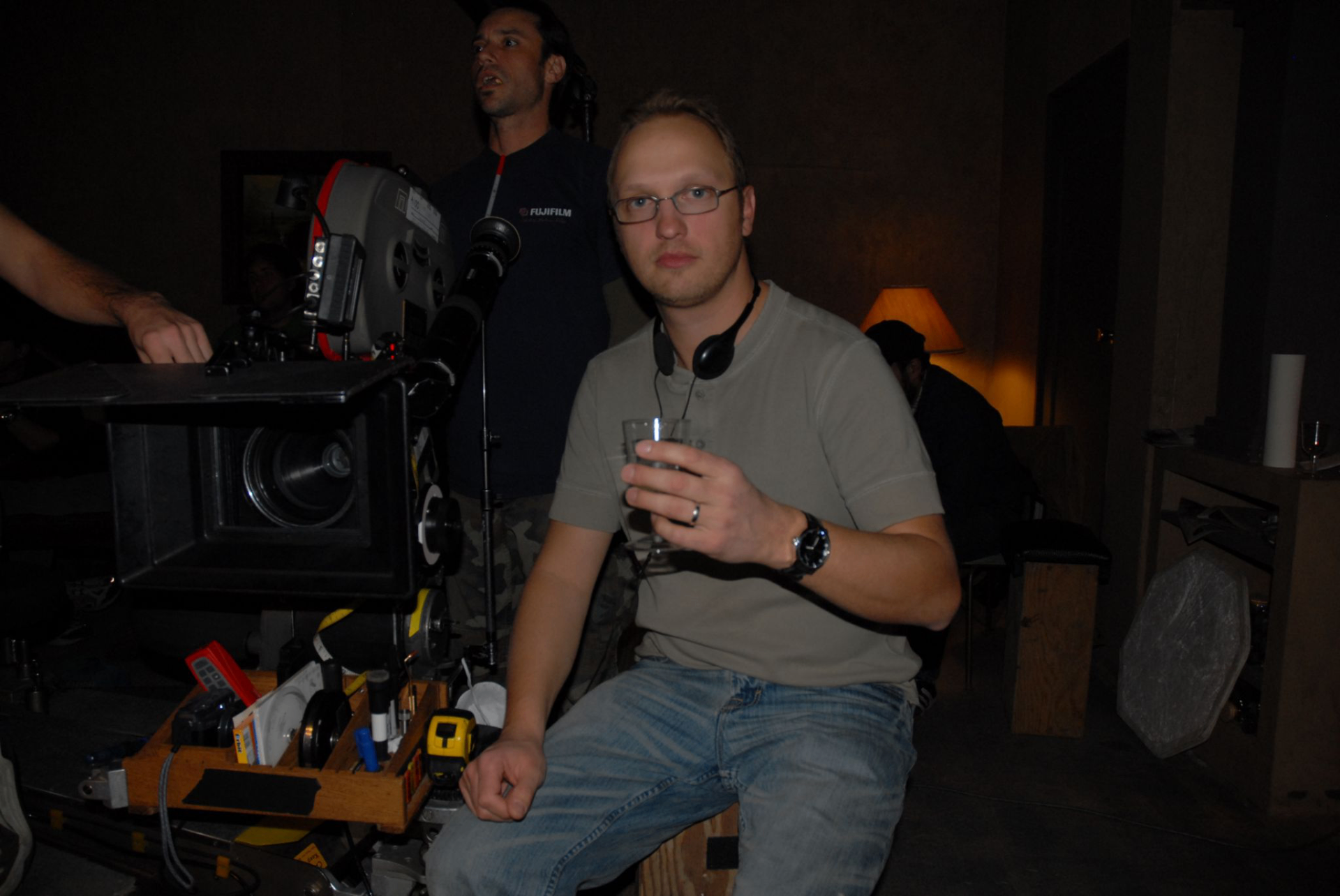 A young filmmaker sitting beside a 35mm film camera on a dimly lit set, holding a glass of water with crew working in the background
